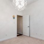 A bright, minimalist children's room with white walls, beige carpet, a decorative chandelier, animal wall décor, and a white bookshelf filled with toys and books. An open door leads to a hallway.