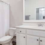 A bright, clean bathroom with white cabinets, a white countertop, a sink with a chrome faucet, a toilet, and a white shower curtain. A large mirror is above the sink, reflecting the open door and hallway.