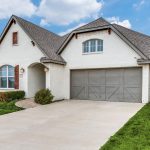 A modern single-story house with white brick exterior, gray double garage doors, gabled roof, arched window, and a wide driveway, surrounded by green grass and neighboring homes under a partly cloudy sky.