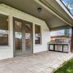 A covered backyard patio with light-colored brick walls, double glass doors with blinds, a large stainless steel grill, tiled flooring, and a wooden fence, overlooking a green lawn and trees under a blue sky.