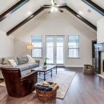 A bright living room with vaulted ceiling, exposed beams, gray sofa, brown leather chair, fireplace, large windows, wooden floor, indoor plant, and a basket with blankets on a patterned rug.
