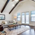 A bright living room with vaulted ceiling, exposed wood beams, a gray sofa, brown armchair, coffee table, large rug, plant, basket, framed art, and French doors letting in natural light.