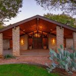Stone house with a prominent wooden entrance, large pillars, and warm exterior lighting. Surrounded by trees, prickly pear cactus, and landscaped gravel, with a green lawn in the foreground.