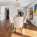 A bright dining area with a round table and four cream chairs on a wooden floor. There’s an antler chandelier, a breakfast bar with patterned stools, large windows, and colorful art on the wall.