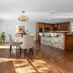 Spacious open kitchen and dining area with wood floors, rustic wood cabinets, stone breakfast bar, stainless steel appliances, beige upholstered chairs around a round table, and antler chandelier. Large windows let in natural light.