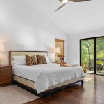 A bright bedroom with a large bed, wooden nightstands, lamps, and leopard print pillows. Sliding glass doors open to a deck with greenery outside, and natural light fills the room through two windows with woven shades.