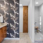 Modern bathroom with a wood vanity, decorative bird-patterned wallpaper, large mirror, towel hanging, potted plant, and a glass-enclosed shower with white tiles and a wooden bench.