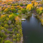 Aerial view of a calm river surrounded by dense trees with autumn foliage, a wooden dock, and a few houses and buildings scattered in the background amidst open fields.