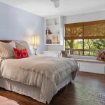 A cozy bedroom with a neatly made bed, two bedside lamps, built-in shelves, a window seat with cushions, and large window with bamboo shades overlooking green trees. The room has wood floors and soft lighting.