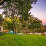 A cozy wooden porch overlooks a lush green lawn, swimming pool, and a serene river surrounded by trees at sunset, with a colorful sky in the background.