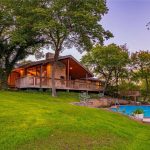 A modern house with a large deck surrounded by trees sits on a grassy hill overlooking an outdoor swimming pool, with outdoor seating and evening lighting creating a cozy atmosphere.