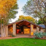 A modern ranch-style house with stone walls, large windows, and a prominent sloped roof, surrounded by green grass, cactus plants, and tall trees in warm, golden sunlight.