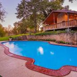 A serene backyard with a clear blue swimming pool bordered by red brick, surrounded by grass and trees, and a stone house with a wooden porch and stairs leading down to the pool area.