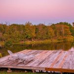 A wooden dock with two lounge chairs and a canoe sits beside a calm river, surrounded by lush trees in early autumn colors under a pastel pink and purple sky at sunset.