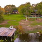 A wooden dock with two lounge chairs sits by the edge of a calm lake, surrounded by green grass and trees. A house and a swimming pool are visible in the background among the trees.