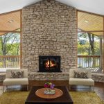 A cozy living room with large stone fireplace, two modern chairs, a coffee table with flowers, and floor-to-ceiling windows showcasing trees and a wooden deck outside.