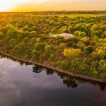 Aerial view of a lush, green tree-covered peninsula surrounded by calm water at sunset, with golden sunlight illuminating the landscape and fields in the background.