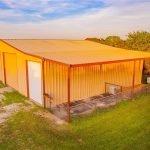 A tan metal barn with a large covered porch sits on a grassy lot, surrounded by trees and under a partly cloudy sky at sunset.