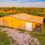 A large metal barn with three closed garage doors sits on a grassy lot, surrounded by trees and fields under a clear blue sky at sunset. A house is visible in the background among the trees.