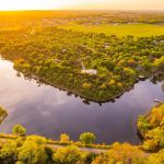 Aerial view of a lake surrounded by lush green trees and scattered houses at sunset, with golden sunlight illuminating the landscape and distant fields.
