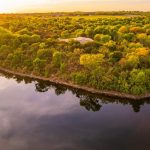 Aerial view of a tree-covered peninsula surrounded by calm water at sunset, with lush green and yellow foliage and rooftops peeking through the trees. The sky is golden, reflecting softly on the water’s surface.