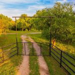 A dirt driveway runs through a metal gate frame and is lined with black fencing, surrounded by green grass and trees under a blue sky with scattered clouds.