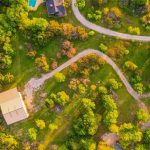 Aerial view of winding roads and driveways through a lush green landscape with scattered trees, a large building, a small house, and a pool near a body of water, all bathed in golden sunlight.