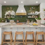 A modern kitchen with a marble island, four wicker stools, white cabinets, and green herringbone tile backsplash. Two large orchid arrangements sit on the island, with stainless steel appliances and open shelving in the background.