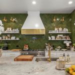 A stylish kitchen with a marble island, two large orchid plants in stone pots, a gold faucet, and a centerpiece of lemons. The back wall features green herringbone tiles, open shelves with dishes, and a white range hood.
