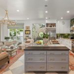 A spacious, elegant kitchen and living area features a large island with drawers, a cozy sitting area by a fireplace, gold accents, green backsplash, modern appliances, and ample natural light from large windows.