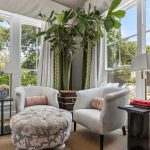 A bright sitting area with two white armchairs, a patterned ottoman, a large indoor tree, and tall windows letting in natural light, overlooking a green garden outside.