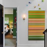 A modern hallway with a striped colorful wall art, bird sculptures above it, two wall sconces, and an open door revealing a green bedroom. On the left, there’s a black door leading to a bathroom with a marble sink.