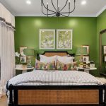 A bedroom with green walls, a double bed with a rattan headboard, striped bedding, and colorful cushions. Two botanical prints hang above the bed, and two green lamps sit on round nightstands. A large mirror is on the right wall.