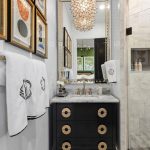 A stylish bathroom with marble walls, a glass shower, a black vanity with gold handles, framed art on the wall, white monogrammed towels, and a decorative chandelier above the sink.