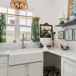 Bright laundry room with white cabinets, a farmhouse sink, marble countertops, patterned floor tiles, tropical curtains, a wicker pendant light, and colorful wall art and decor on shelves and walls.