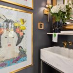 A stylish bathroom features a marble sink, gray walls, gold accents, a large framed portrait of a woman with flowers in her hair, white hydrangeas in a vase, and a wall-mounted faucet beneath a shelf with decor.