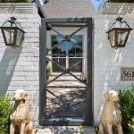 A metal gate framed by white brick walls with two lantern-style lights and a house number plaque reading 5614. Two dog statues sit on either side of the entrance, surrounded by green bushes and yellow flowers.