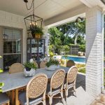 A covered patio with a dining table and wicker chairs overlooks a backyard with a pool, lounge chairs, green lawn, and trees. Potted plants and decorative items adorn the table and shelves.