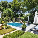 A backyard with a rectangular swimming pool, surrounded by lounge chairs, umbrellas, plants, and seating areas. Trees and a wooden fence line the backyard, creating a private, lush outdoor space.