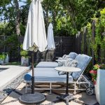 Three light-blue cushioned lounge chairs with white umbrellas are arranged beside a pool, surrounded by greenery, tall planters, and a black wooden fence covered in leafy vines.