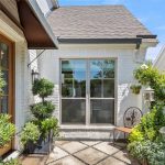 Sunny courtyard with white brick walls, large window, green topiary plants, a wooden bench, potted greenery, and trellis vines climbing along the wall near double wooden doors.