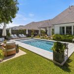 A backyard with a rectangular swimming pool, surrounded by lounge chairs, outdoor seating, potted plants, and a grassy lawn, next to a light-colored house under a clear blue sky.
