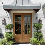 A double wooden door with glass panels is set in a white brick wall under a metal awning. Two large potted plants flank the entrance, and wall-mounted lanterns are on either side. A welcome mat lies on the stone walkway.