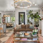 Bright, stylish living room with a brown sofa, glass coffee table, and fireplace with a TV above. Open kitchen with bar stools, dining table, large plants, and ample natural light from big windows.