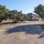 A gravel driveway curves toward a stone house with a steep roof and a visible American flag. The entrance is flanked by two large rocks marked with house numbers and lined with trees.