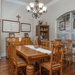 A traditional dining room with a wooden table and chairs, a lace table runner, wooden cabinets displaying decorative plates and vases, a chandelier, and a window showing trees and brickwork outside.