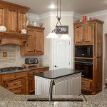 A spacious kitchen with wooden cabinets, granite countertops, a small island, built-in oven and microwave, a gas cooktop, and decorative items on top of the cabinets. Pendant lights hang above the island.