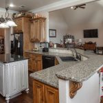 Spacious kitchen with wooden cabinets, granite countertops, a black fridge, white island, and built-in oven. The kitchen opens to a living area with hardwood floors, fireplace, and a mounted TV. Natural light enters from a glass door.