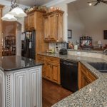 A spacious kitchen with wooden cabinets, granite countertops, a black double sink, a kitchen island with white cabinetry, black appliances, and two pendant lights hanging over the island.