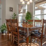 A bright dining area with a wooden table and six chairs, surrounded by large windows and several green potted plants on a hardwood floor. Sunlight fills the room, giving it a warm and inviting atmosphere.
